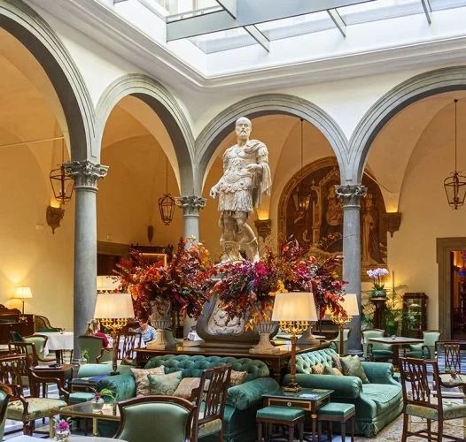 The interior courtyard of the Palazzo Portinari Salviati in Florence, Italy, which has been converted into a luxurious lounge or hotel lobby. The space features grand Renaissance architecture with large arches and columns. A central statue of a Roman figure is surrounded by elaborate floral arrangements.